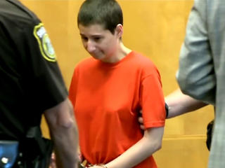 A woman wearing an orange shirt is led out of a courtroom by officers, indicating a legal proceeding image